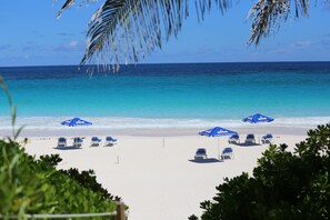 A beach with white sand, clear blue water, and several blue umbrellas. - Tingum Village Hotel (Dunmore Town)
