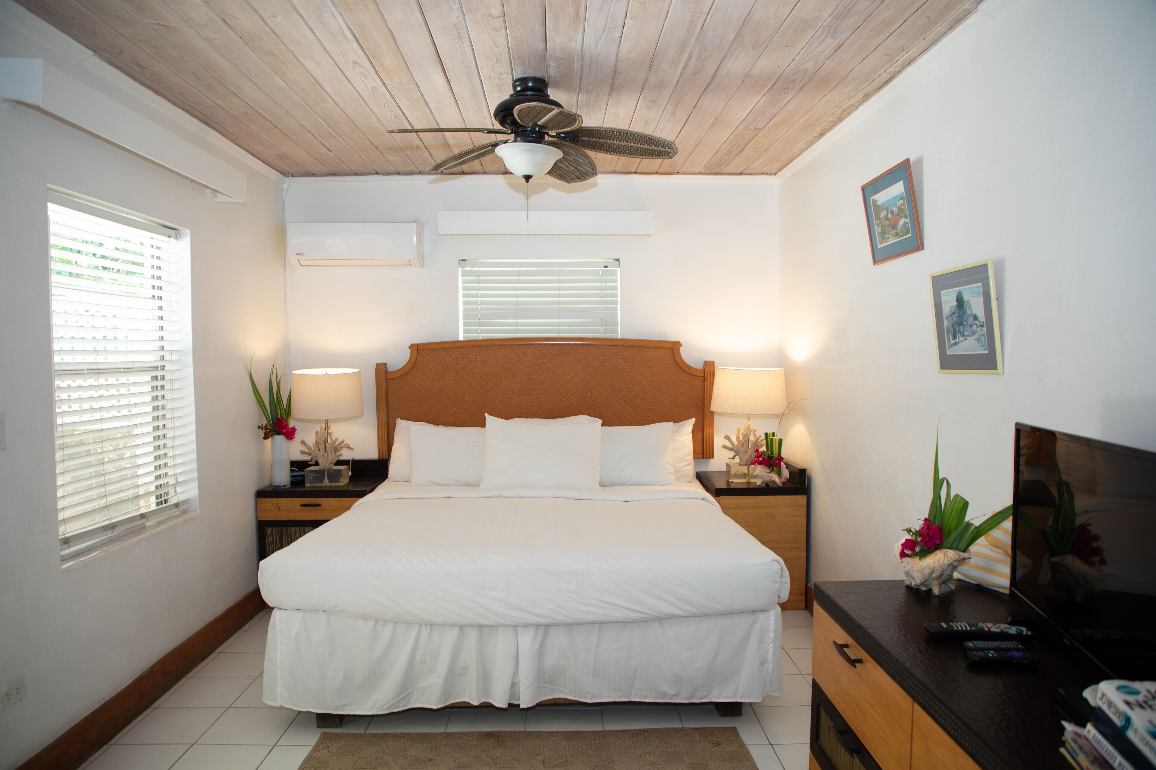 A neatly arranged bedroom with a large bed, wooden ceiling, two bedside tables with lamps, and a TV on a wooden dresser.
