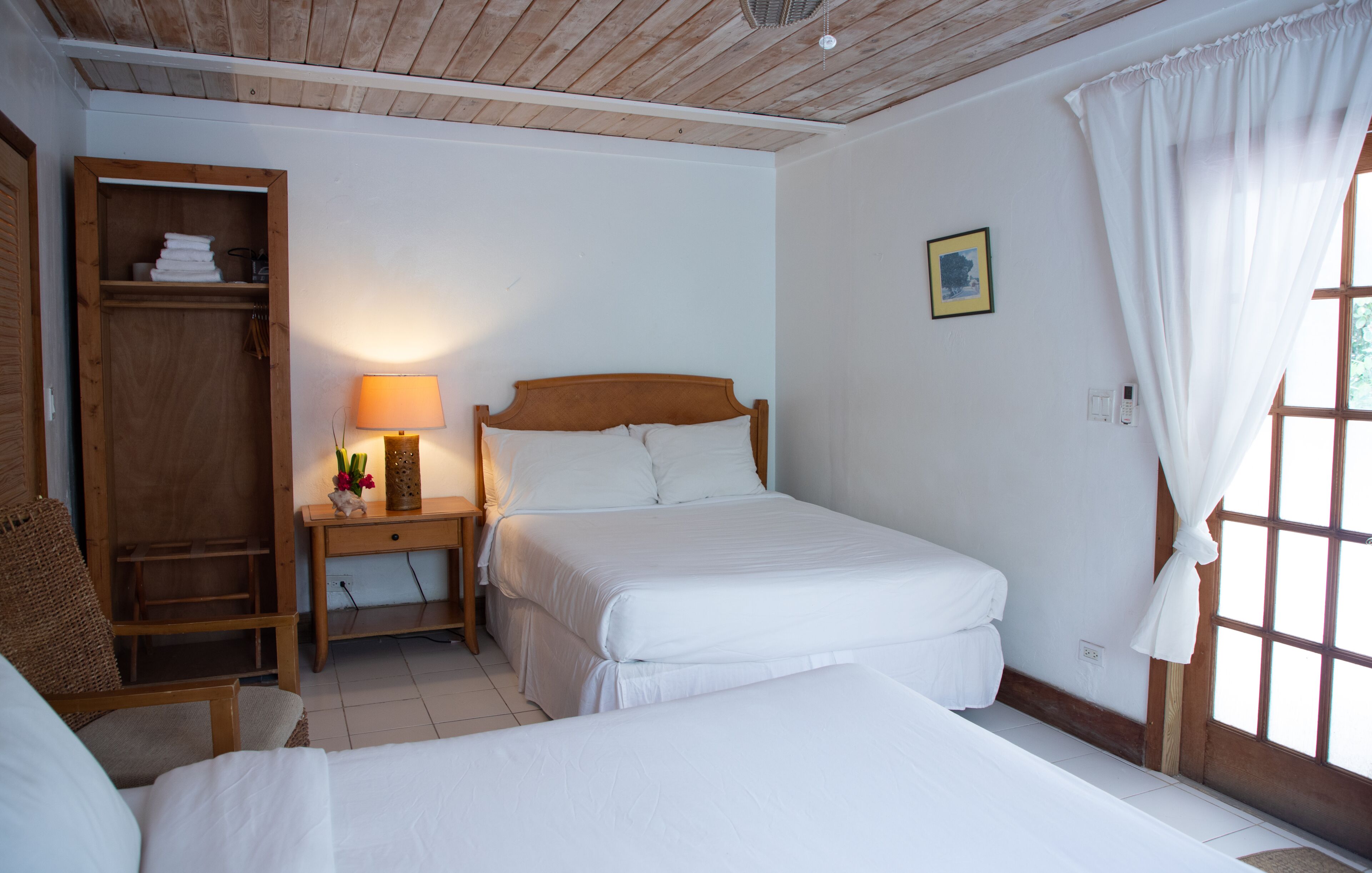 A room with two beds, a wooden wardrobe, a bedside table with a lamp, and a window with curtains.