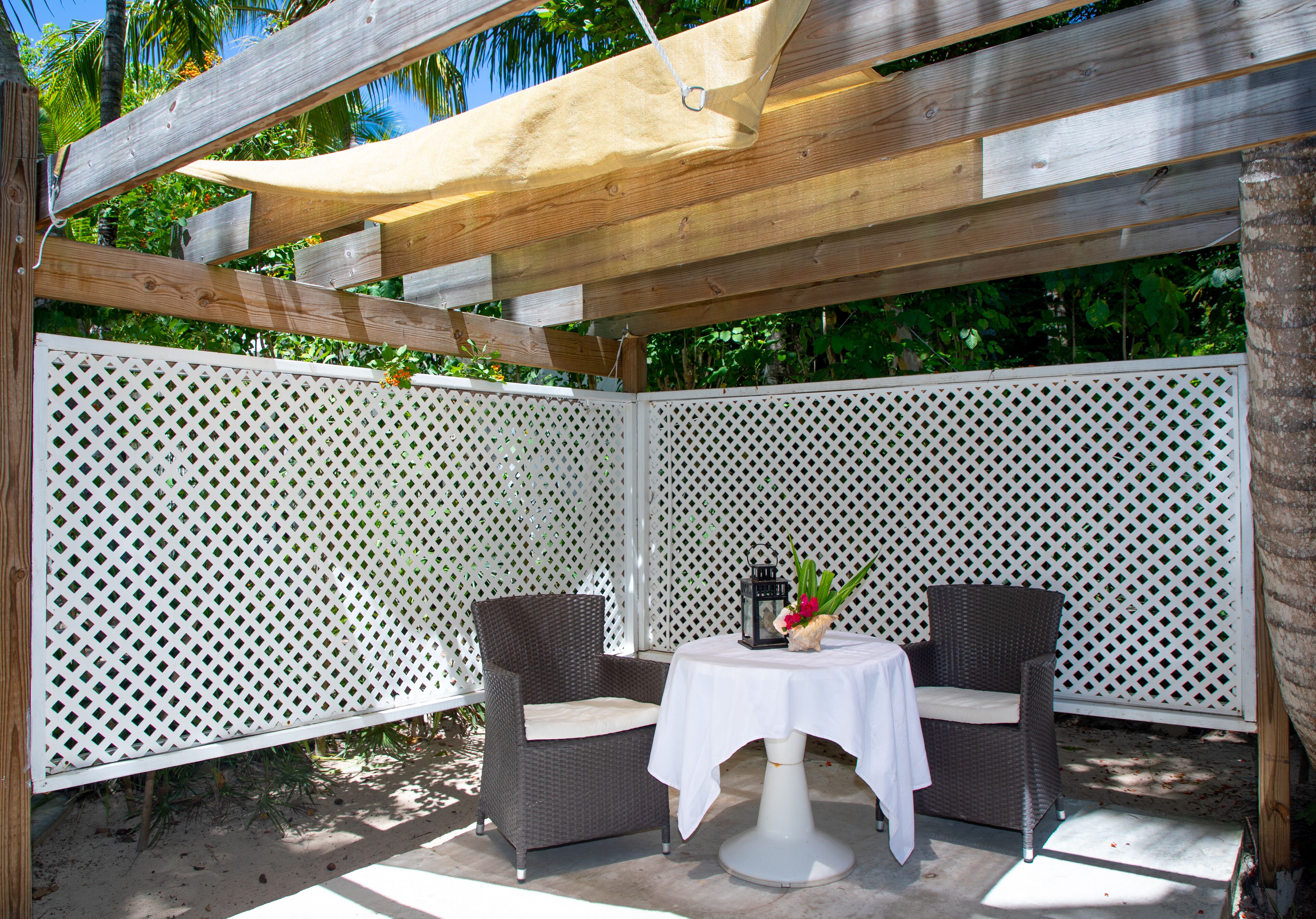En overdækket terrasse med to fletstole og et bord, omgivet af et hvidt espalier.