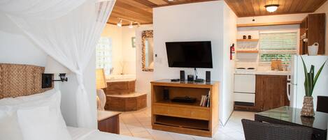 A bedroom with a bed, a TV, a wooden cabinet, and a small kitchen area with a refrigerator and a window.