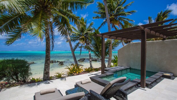 A poolside area with lounge chairs and a pergola overlooking a beach with palm trees and clear blue water.