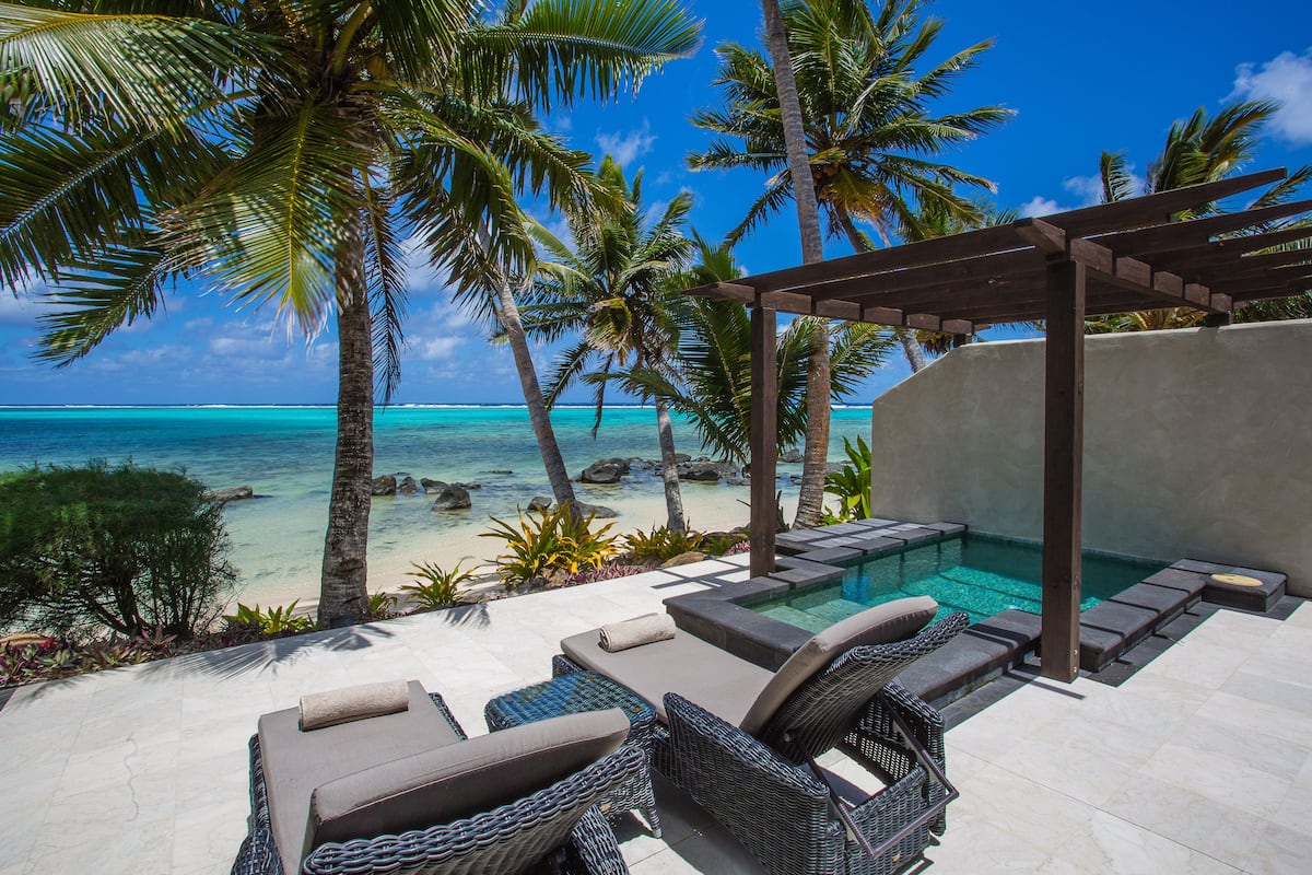 A poolside area with lounge chairs and a pergola overlooking a beach with palm trees and clear blue water.