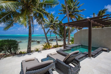 A poolside area with lounge chairs and a pergola overlooking a beach with palm trees and clear blue water.