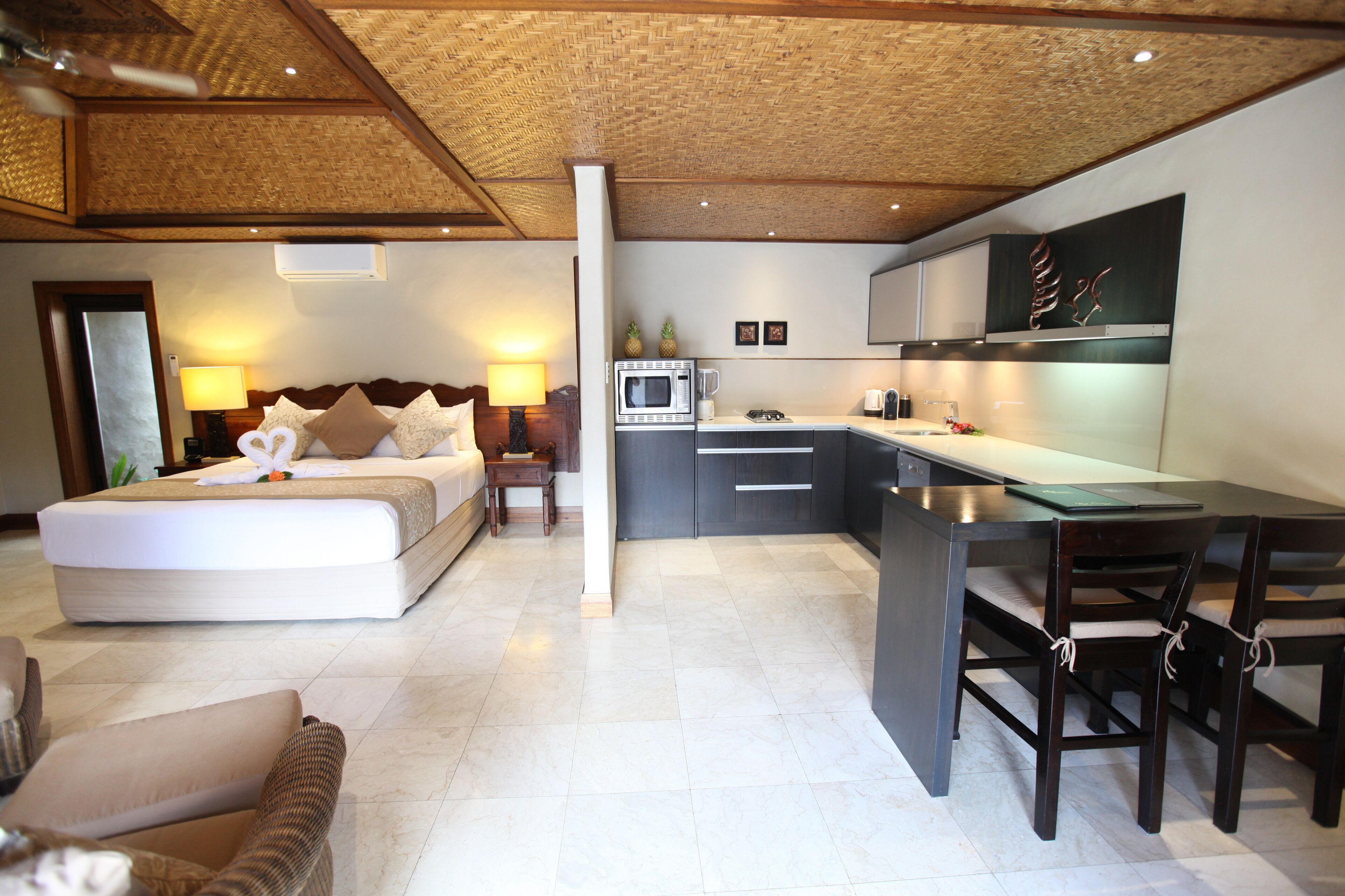 A modern hotel room with a large bed, a sitting area with a sofa, a dining table with chairs, a kitchenette, and a ceiling with a woven design.