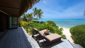 A wooden deck with lounge chairs and a small table, overlooking a beach with palm trees and clear blue water.