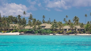 A beachfront property with thatched-roof buildings, palm trees, and clear turquoise water.