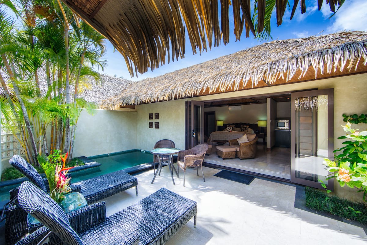 A poolside area with wicker furniture, a thatched roof, and a view into a room with a bed and a TV.