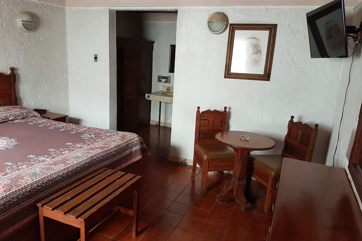 A bedroom with a bed, a wooden bench, a small round table, and a chair. There is a door leading to a bathroom and a wall-mounted TV.