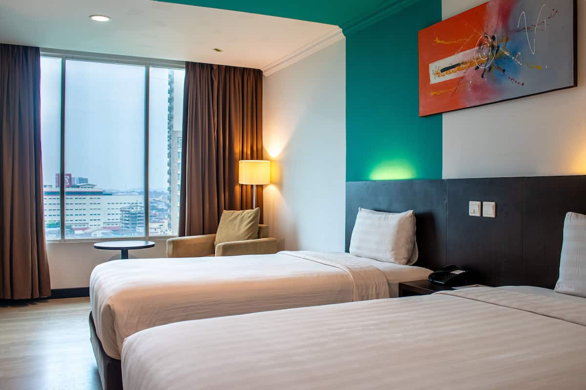 A hotel room with two beds, a view of the city, a small table, and a lamp.