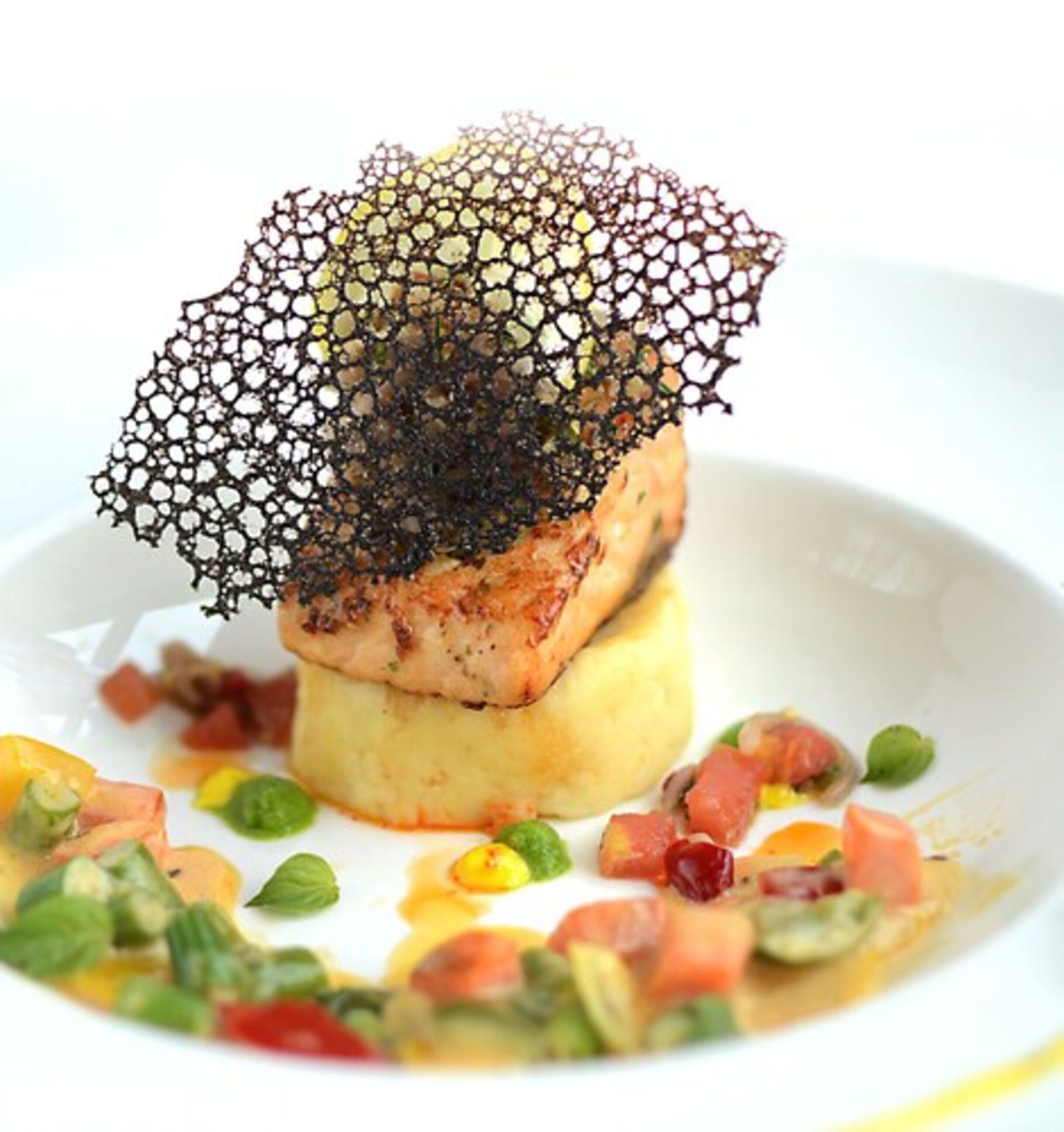 A dish with a fish fillet on a bed of mashed potatoes, garnished with a crispy, web-like structure, and surrounded by a colorful vegetable medley.