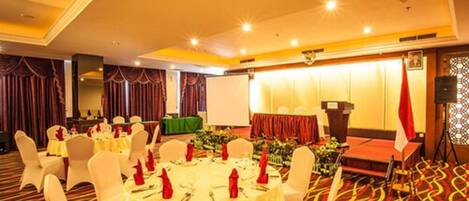 A banquet hall with round tables set for dining, a stage with a podium, and a patterned carpet.