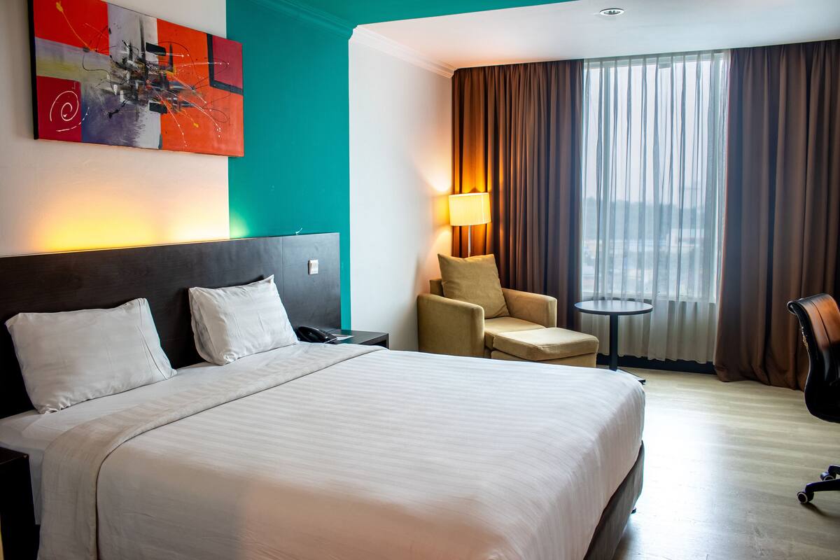 A hotel room with a large bed, a desk, a chair, a lamp, and a view of the city through the window.