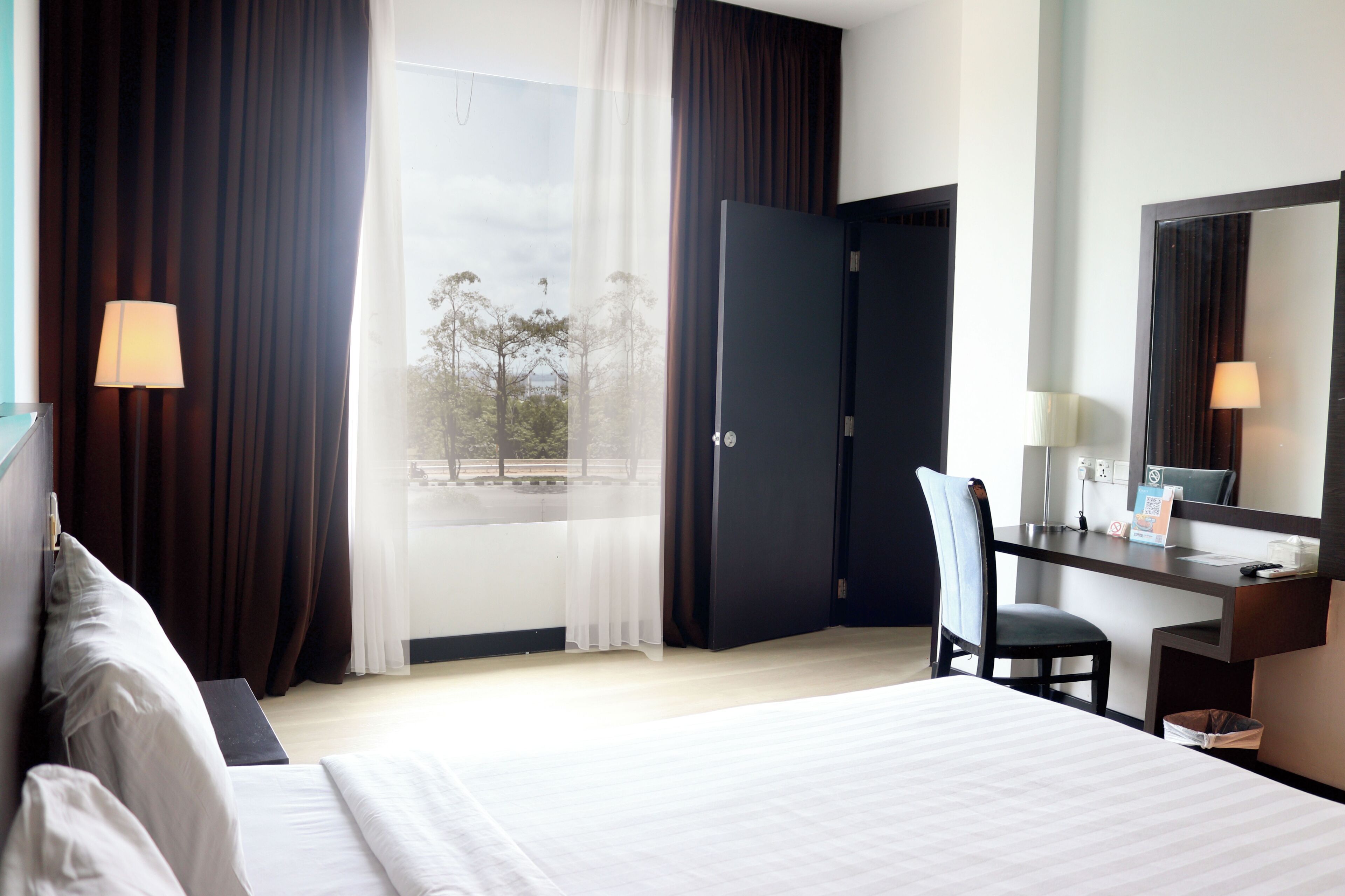 A hotel room with a bed, a desk, a chair, a large window with curtains, and a mirror.