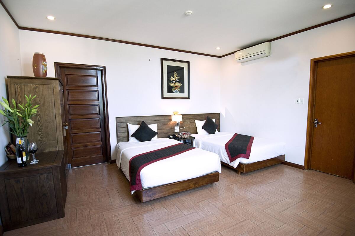 A hotel room with two beds, a wooden wardrobe, a nightstand with a lamp, and a framed picture on the wall.