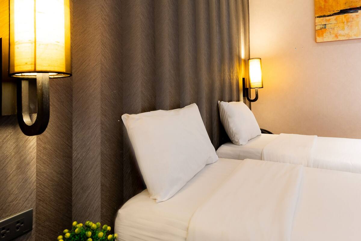 A hotel room with two beds, a nightstand with a lamp, a potted plant, and a painting on the wall.