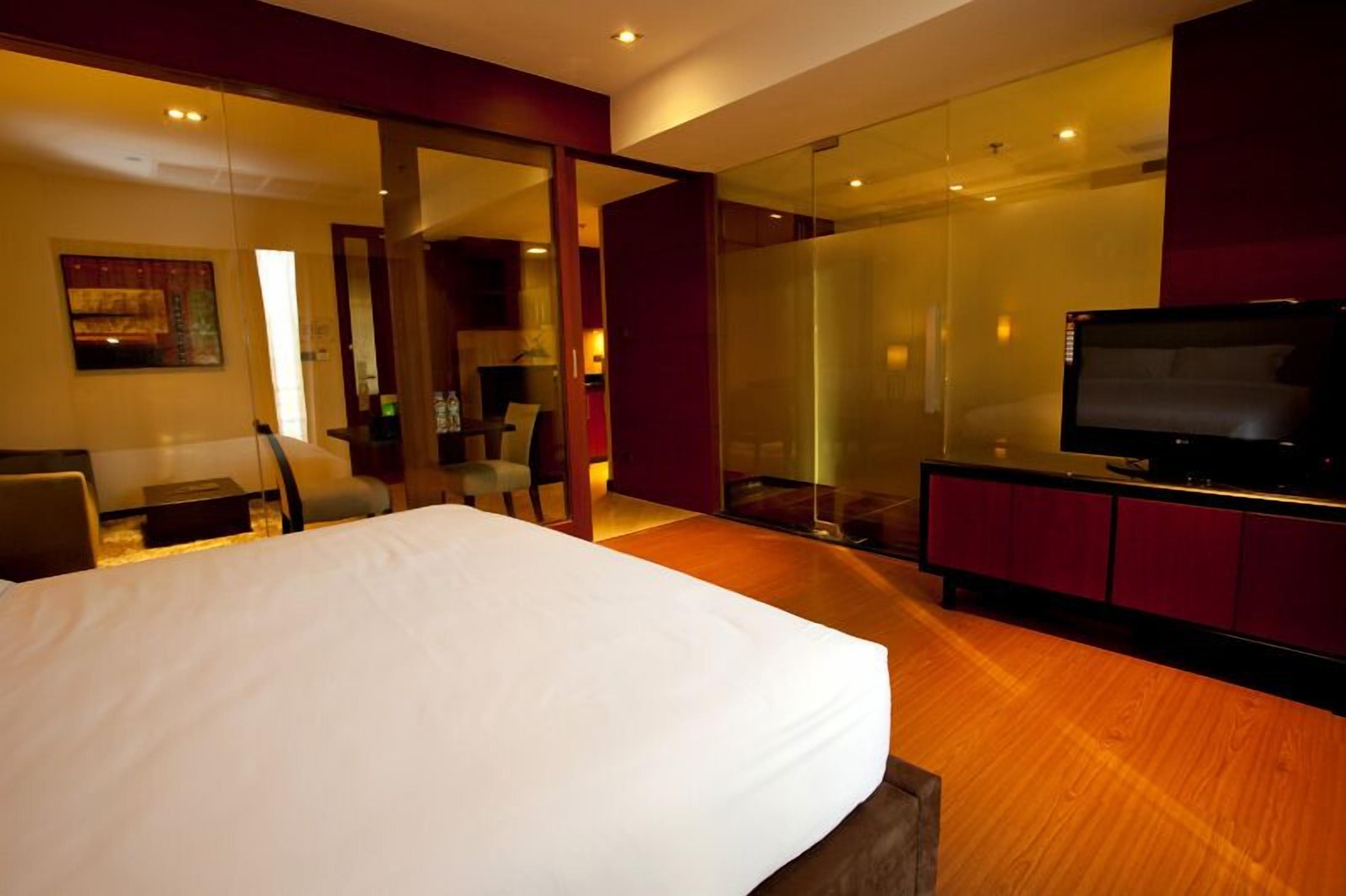Een moderne hotelkamer met een groot bed, een televisie en een glazen scheiding tussen de zithoek en de slaapkamer.