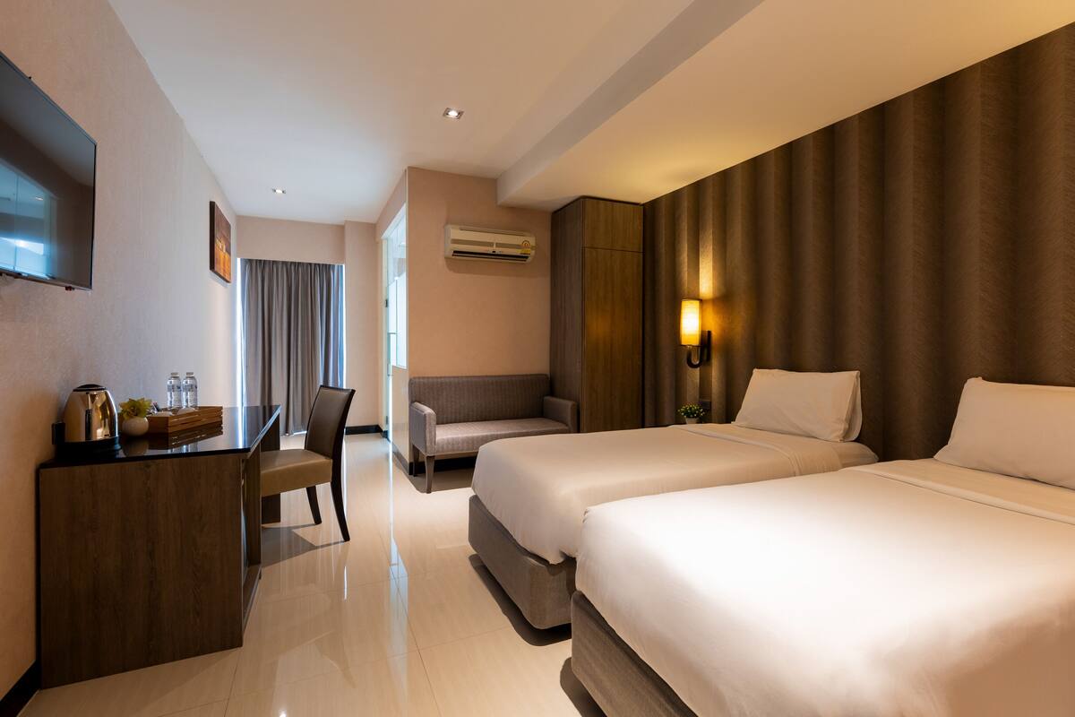 A hotel room with two beds, a desk, a chair, a TV, and an air conditioning unit.