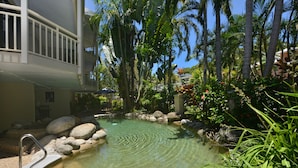 A clear pool surrounded by rocks and tropical plants, with a balcony overlooking the pool area. - Driftwood Mantaray (Port Douglas)