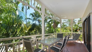 A covered outdoor patio with a glass table and four chairs, surrounded by lush tropical plants and palm trees. - Driftwood Mantaray (Port Douglas)