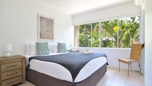 A bedroom with a large bed, a nightstand, a chair, and a window with a view of palm trees. - Driftwood Mantaray (Port Douglas)