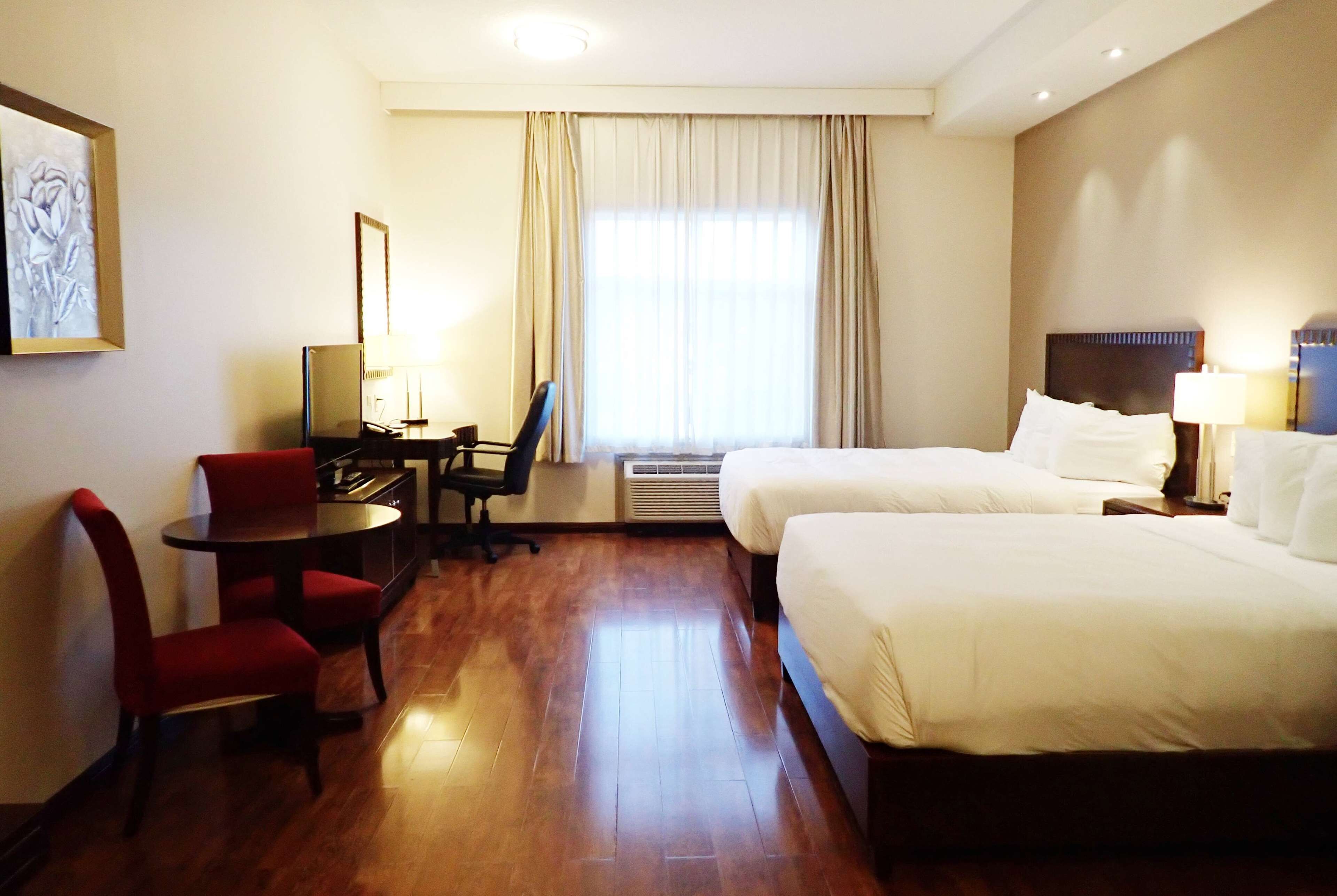 Une chambre d’hôtel avec deux lits, un bureau avec une chaise, une petite table et une grande fenêtre avec des rideaux.