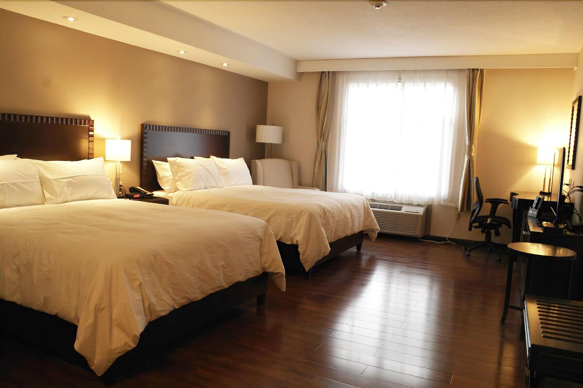 A hotel room with two beds, a desk, an air conditioning unit, and a window with curtains.