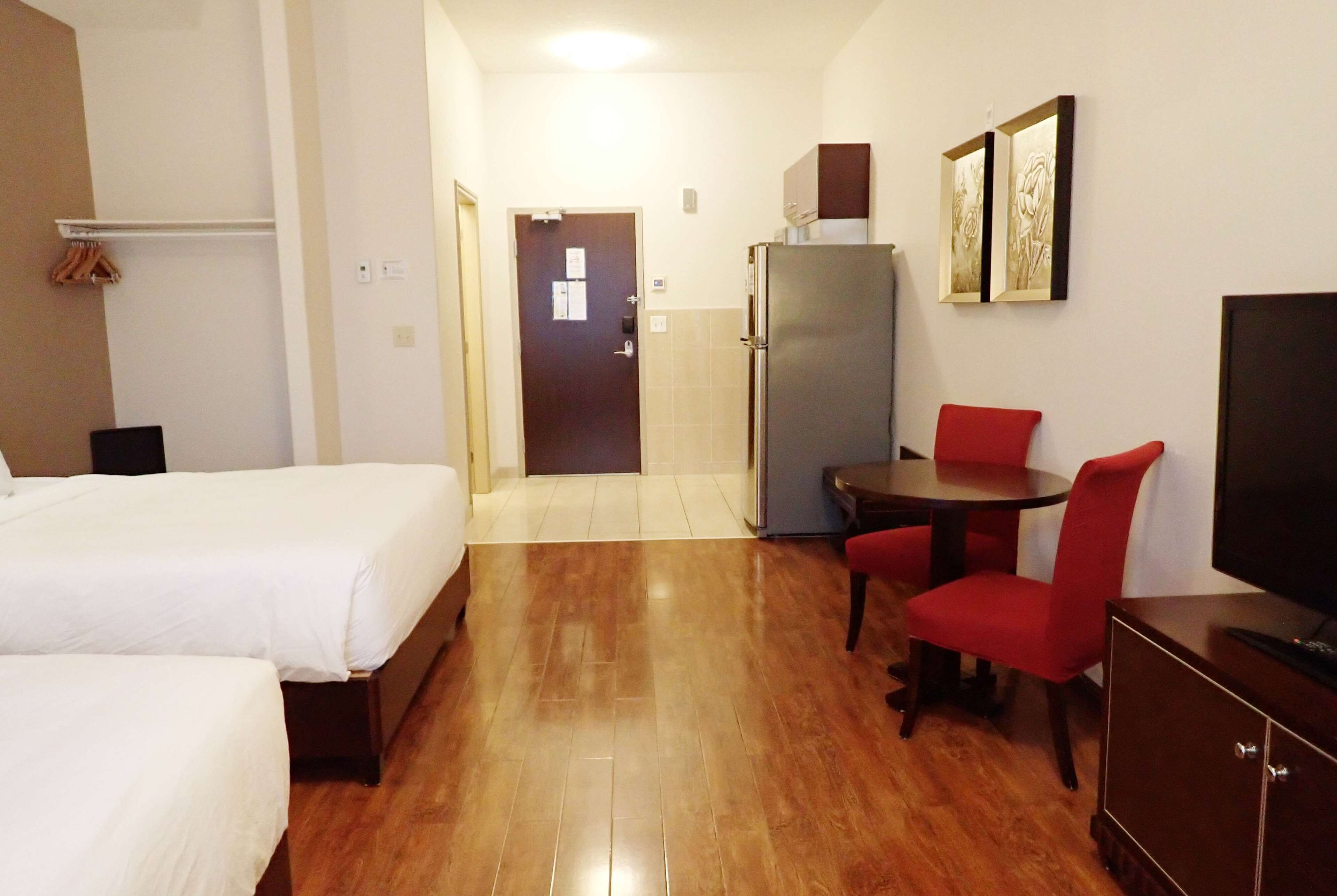 Una habitación de hotel con dos camas, una mesa de comedor con sillas rojas, un refrigerador y un televisor.