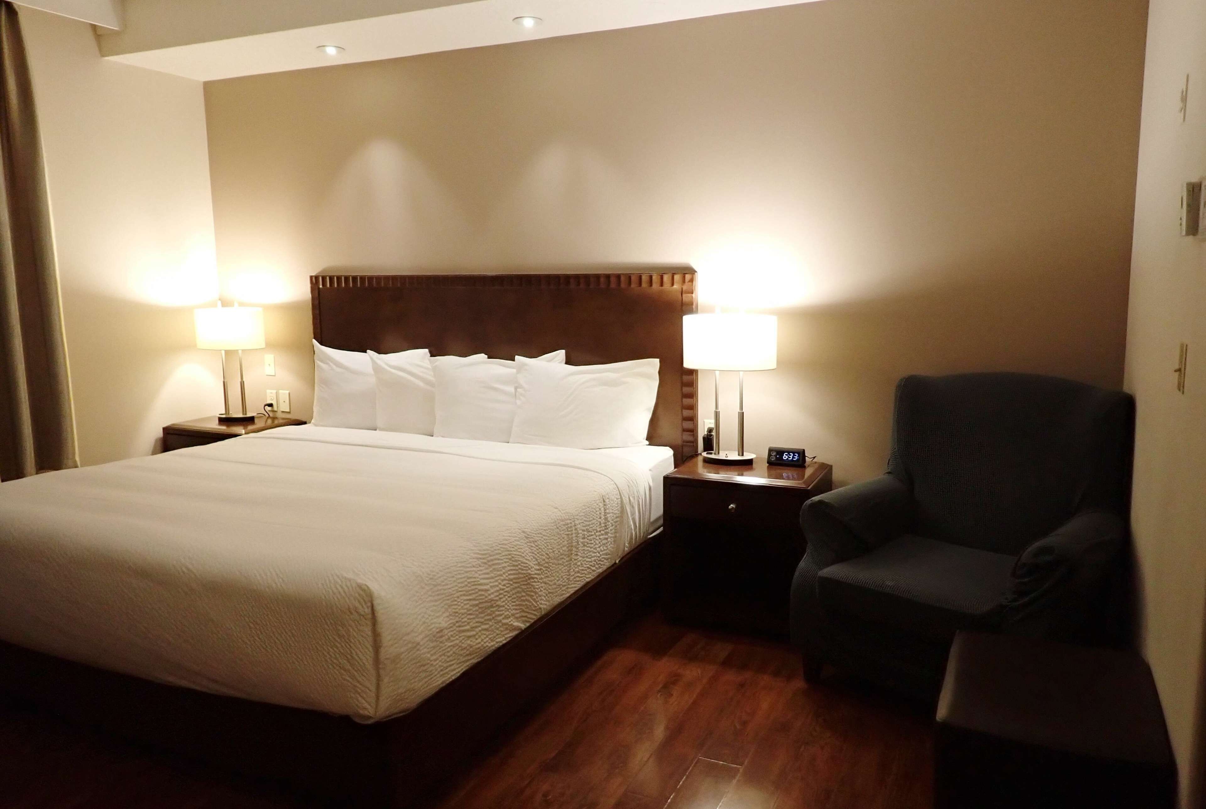 A hotel room with a large bed, two bedside lamps, a chair, and a nightstand with a clock.