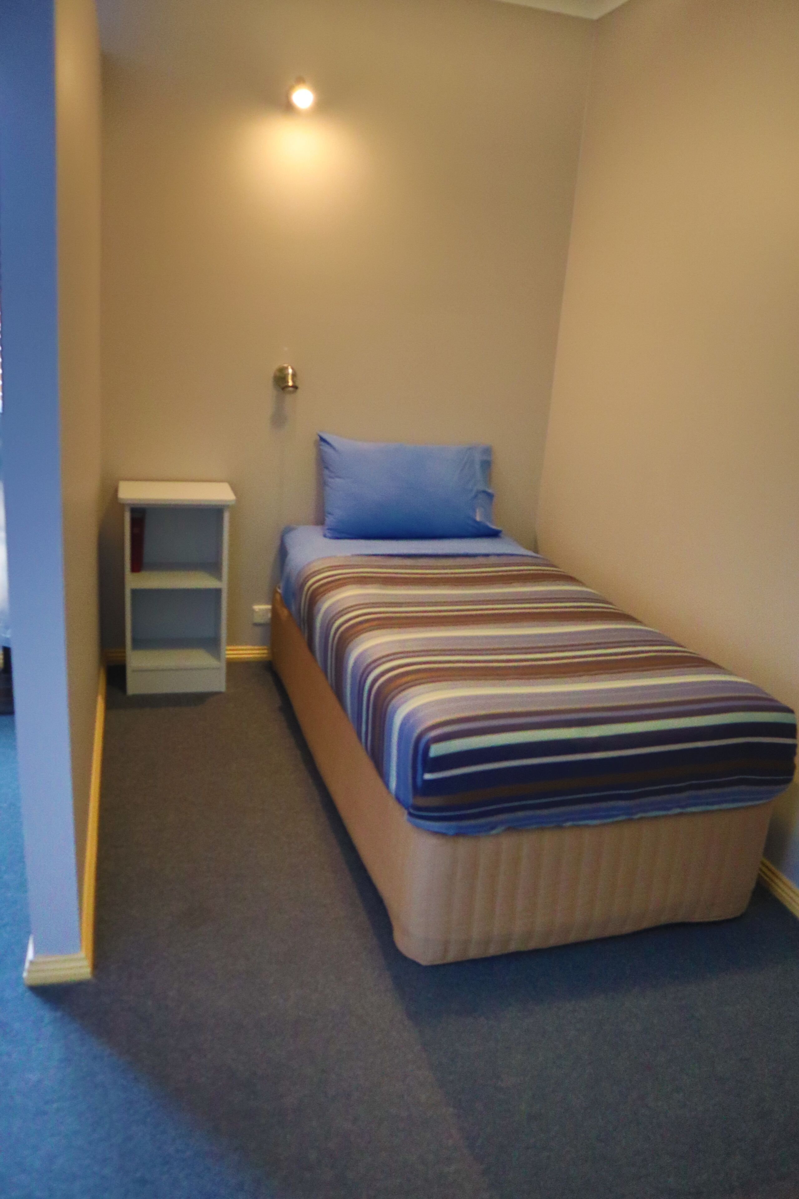 A small, single-bed room with a striped bedspread, a nightstand, and a wall-mounted shelf.
