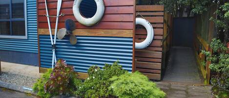 A wooden fence with a blue corrugated metal wall, a wooden door, and a small garden with green plants and a sign that reads "SANDWICH ISLAND".