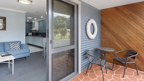 A modern living area with a sofa, a coffee table, and a dining table with chairs. There is a sliding glass door leading to a patio with a wooden wall and a metal chair. - Strahan Bungalows (Strahan)