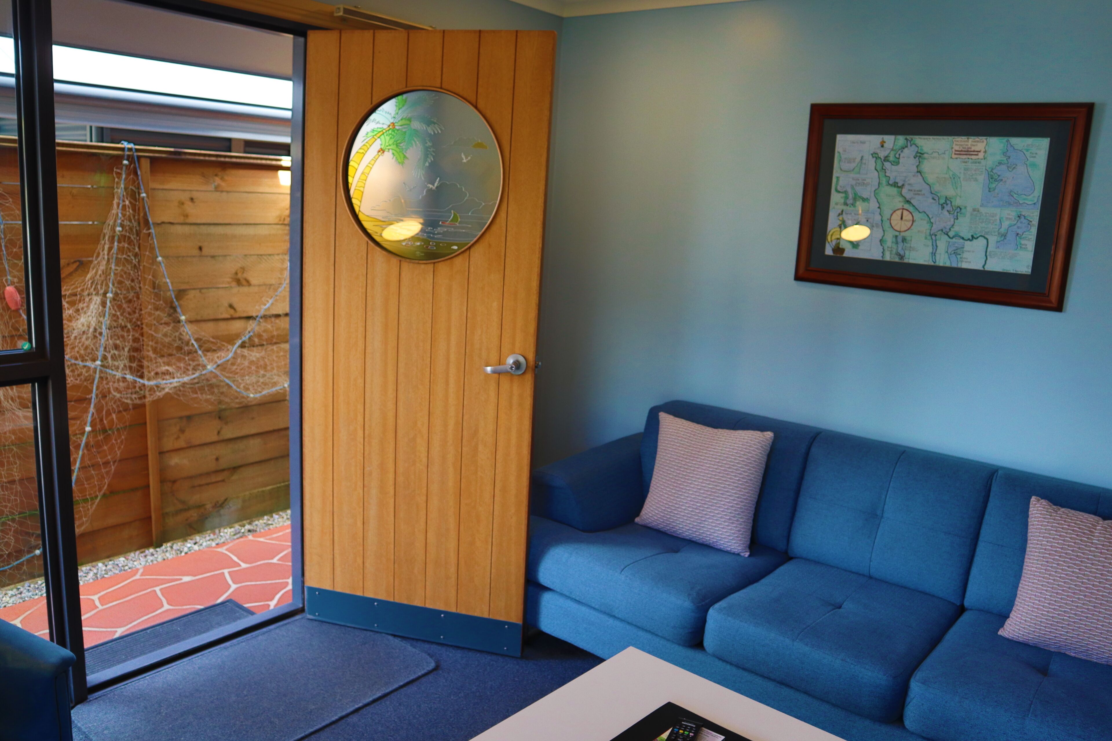 A room with a wooden door, a blue sofa, a map on the wall, and a window with a view of a wooden fence and a net.