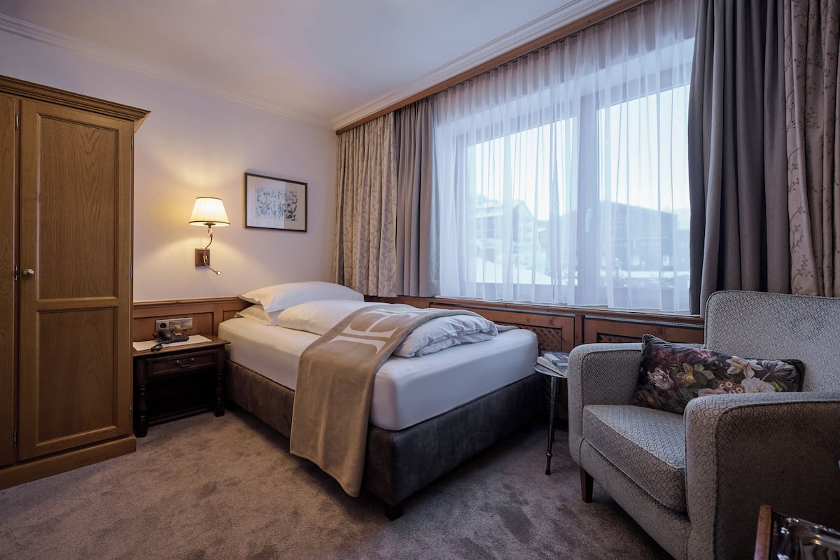 A hotel room with a bed, a nightstand, a chair, a window with curtains, a wall-mounted lamp, and a framed picture.