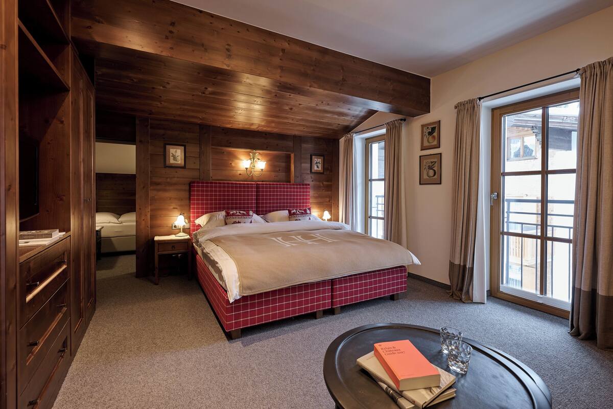 A bedroom with a wooden ceiling, a large bed with a red and white checkered bedspread, a bedside table with a lamp, and a window with curtains.