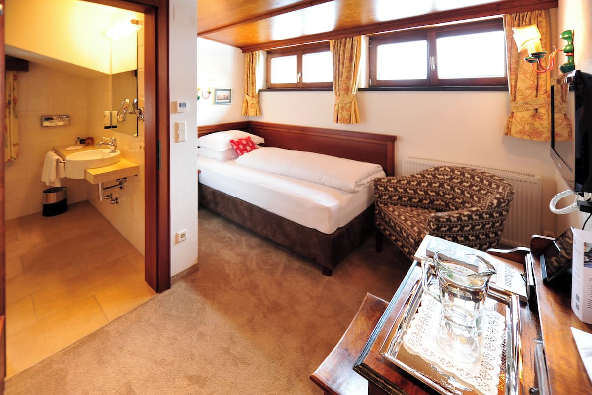 A small, cozy room with a single bed, a bedside table, a chair, a window with curtains, a radiator, and a bathroom with a sink and mirror.
