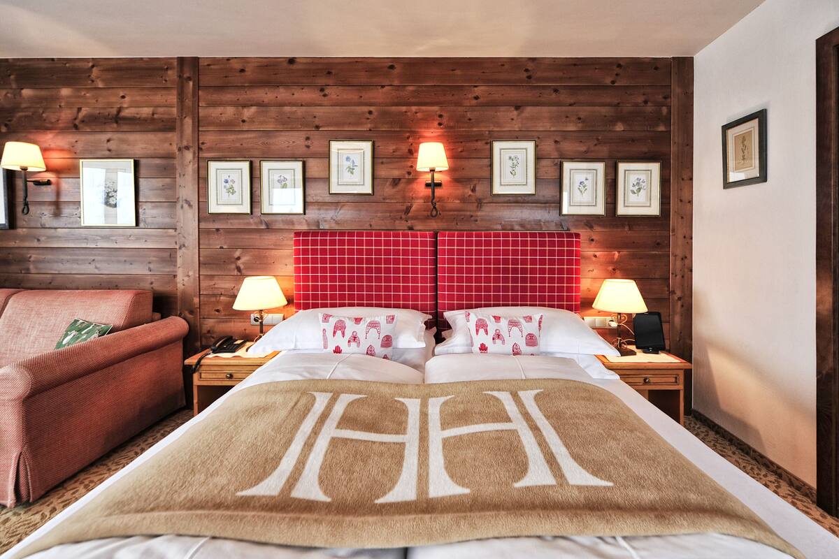 A hotel room with a wooden wall, a red checkered headboard, a large bed with a blanket, two bedside tables with lamps, and a red sofa.