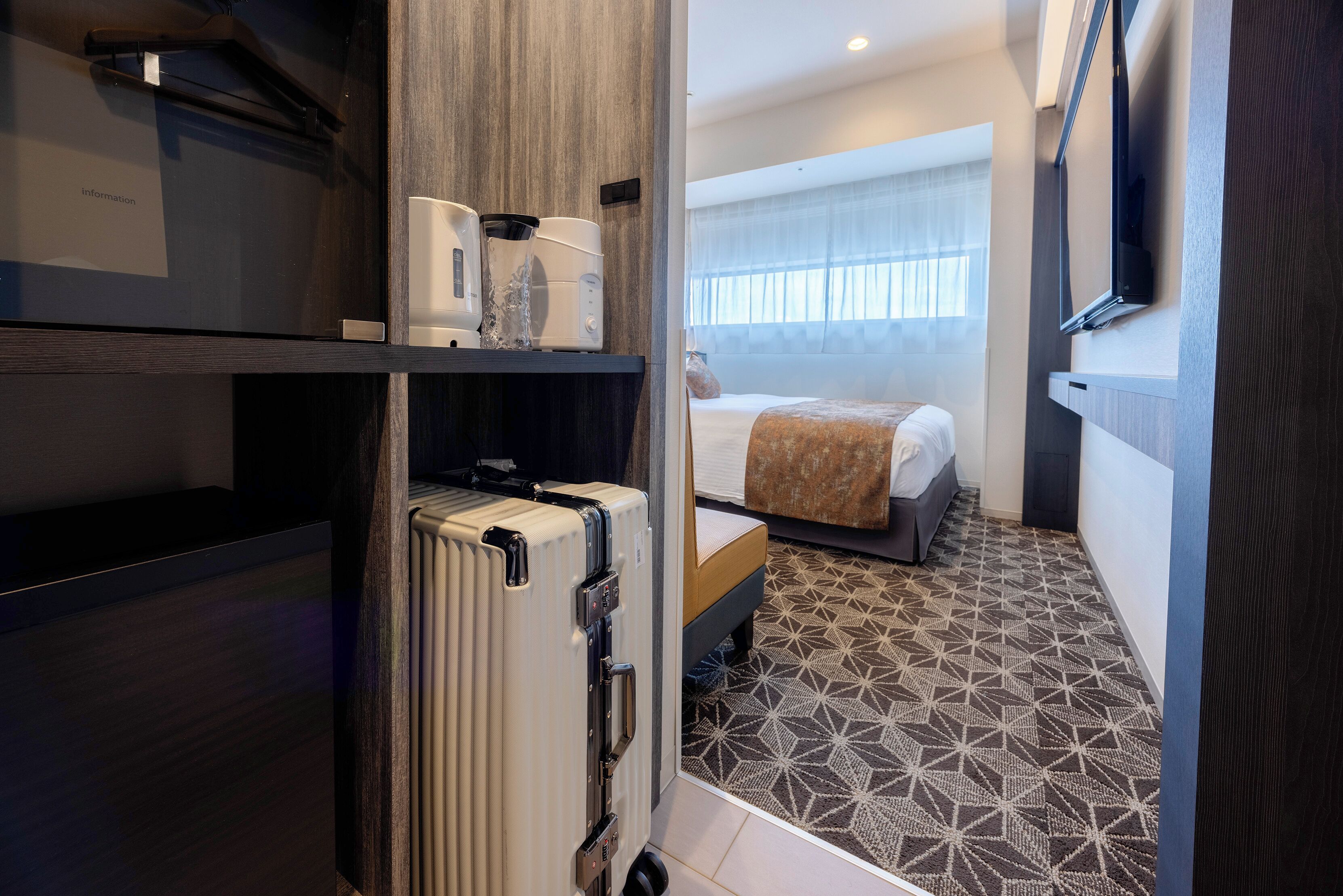 A hotel room with a bed, a radiator, a shelf with a coffee maker, and a patterned floor.