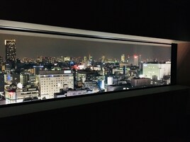 Vista nocturna de un paisaje urbano a través de una ventana, con varios edificios y ventanas iluminadas.