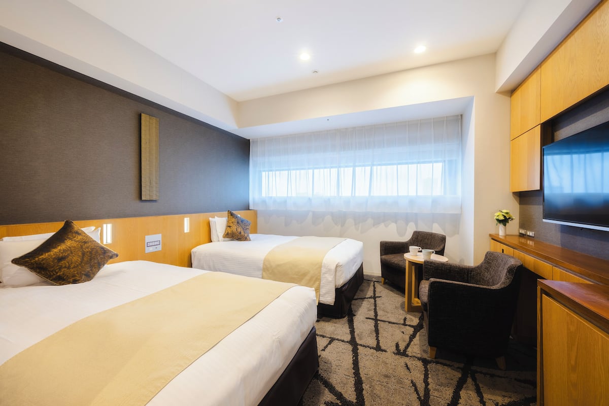 A hotel room with two beds, a small table, a chair, a TV, and a window with sheer curtains.