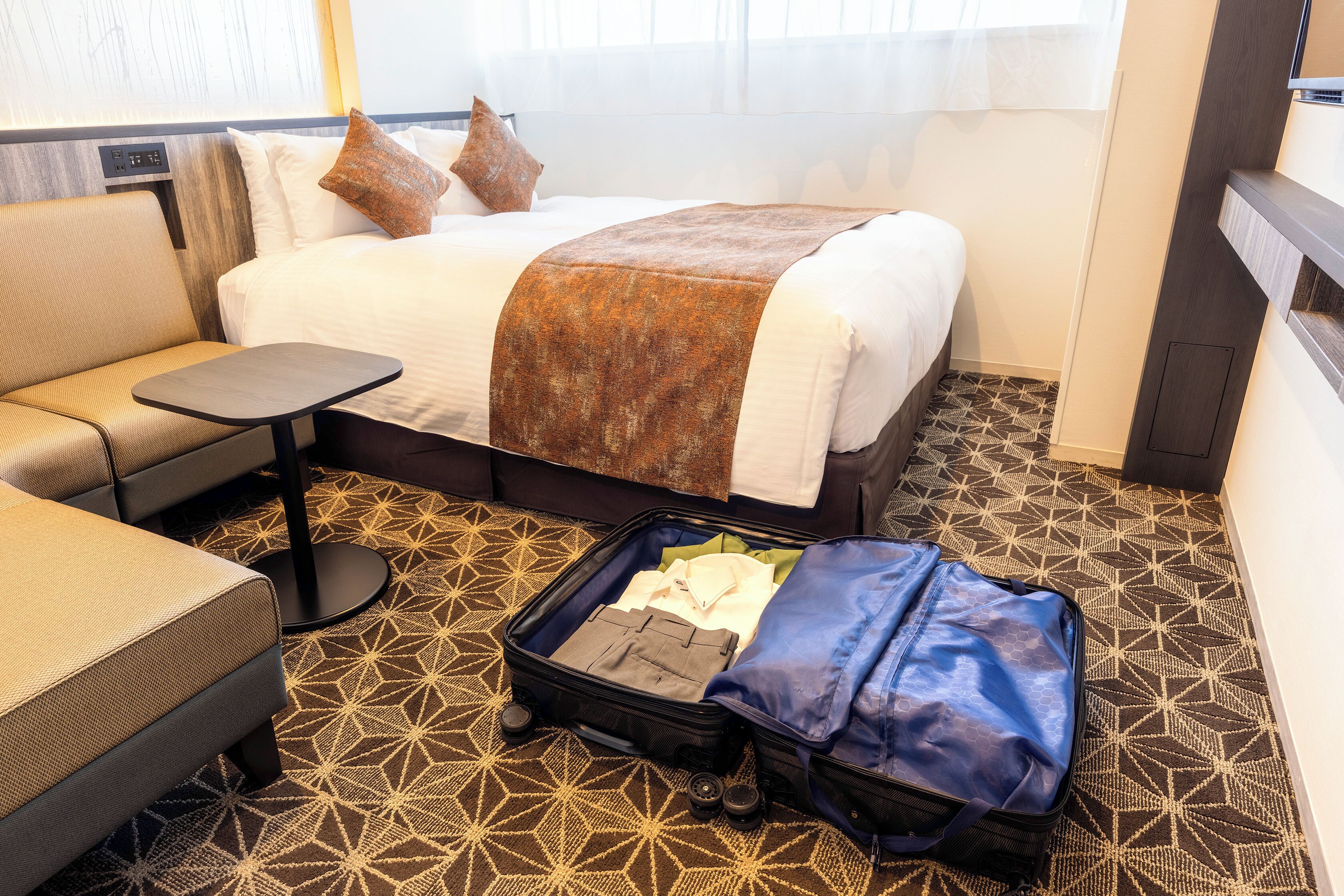 Quarto de hotel com uma cama, uma pequena mesa, uma cadeira e uma mala no chão.