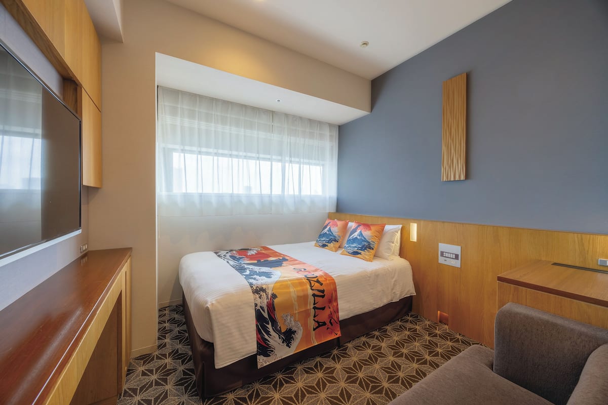 A hotel room with a bed, a TV, a desk, and a window with sheer curtains.
