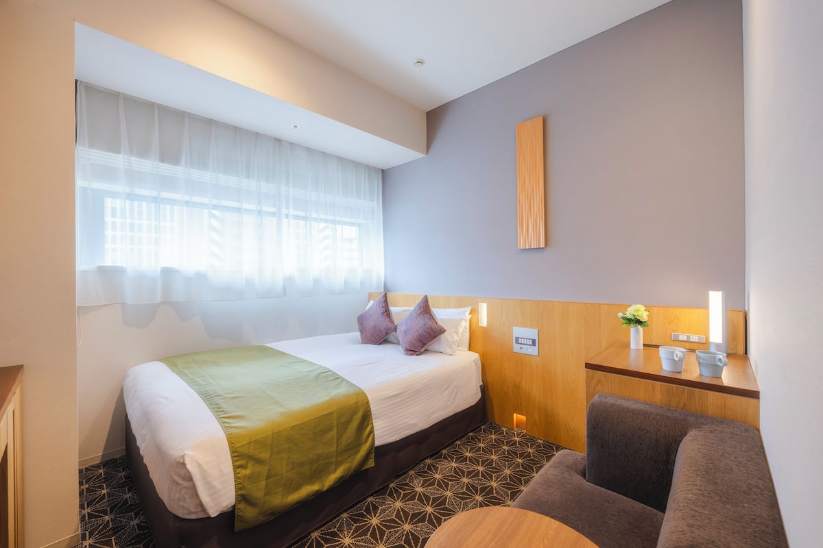 A hotel room with a bed, a desk, a chair, and a window with sheer curtains.