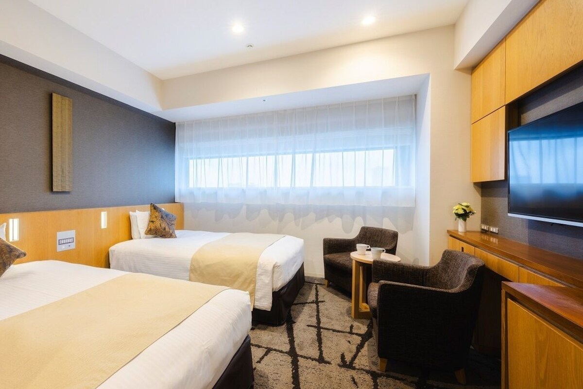 Habitación de hotel con dos camas, un escritorio, una silla, un televisor y una ventana con cortinas translúcidas.