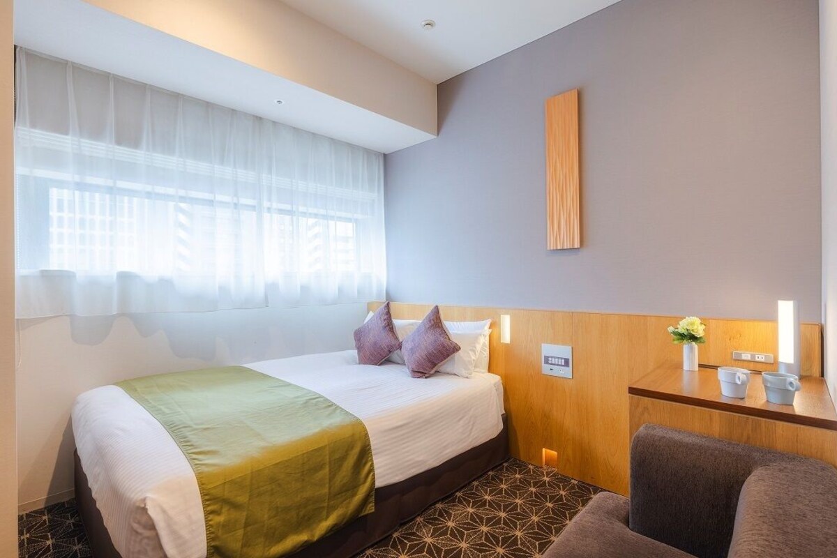Una habitación de hotel con cama, mesita de noche con lámpara, una silla y ventana con cortinas ligeras.