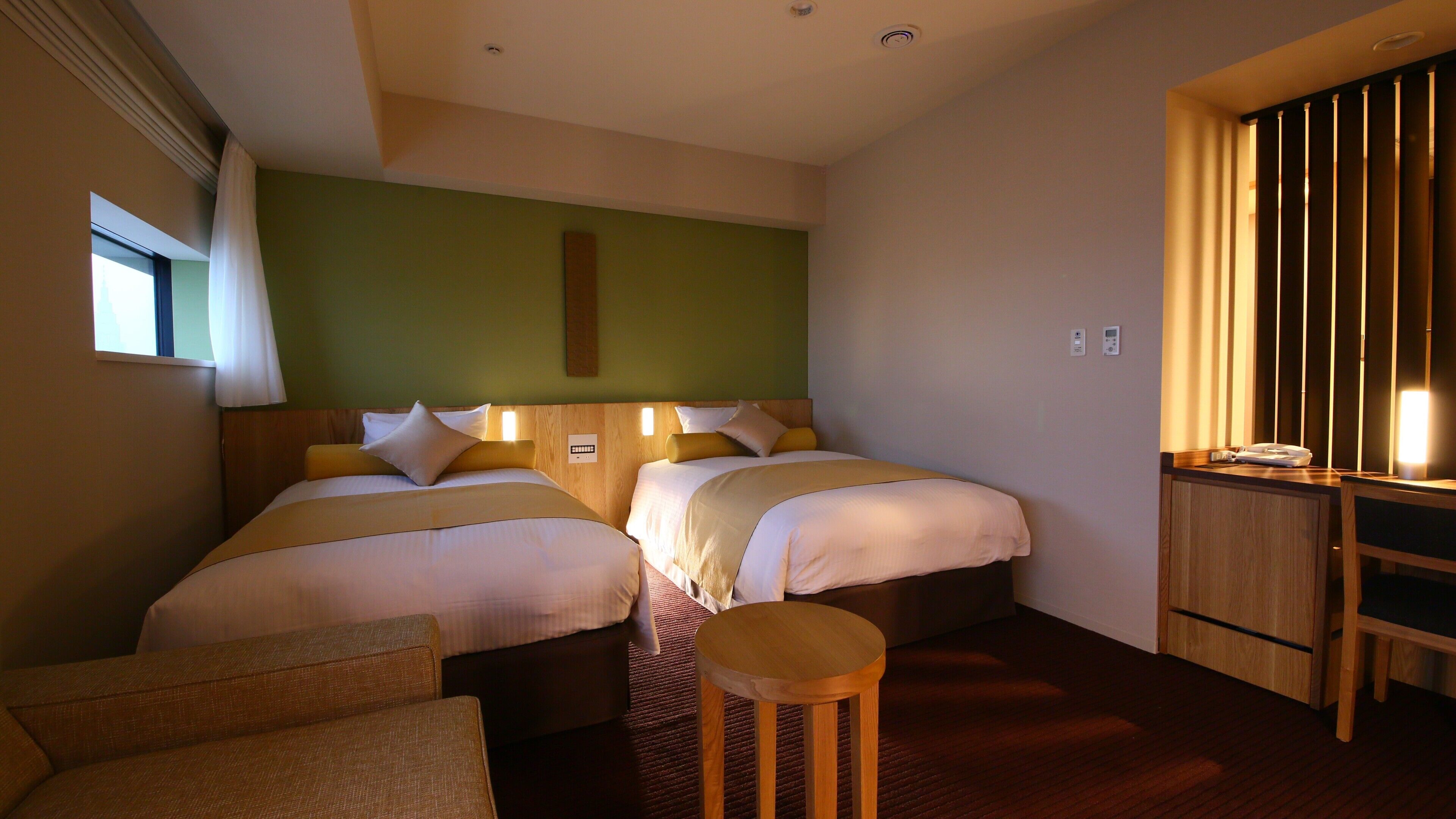 Ein Hotelzimmer mit zwei Betten, einem Schreibtisch, einem Stuhl, einem kleinen Tisch und einem Fenster mit Vorhängen.
