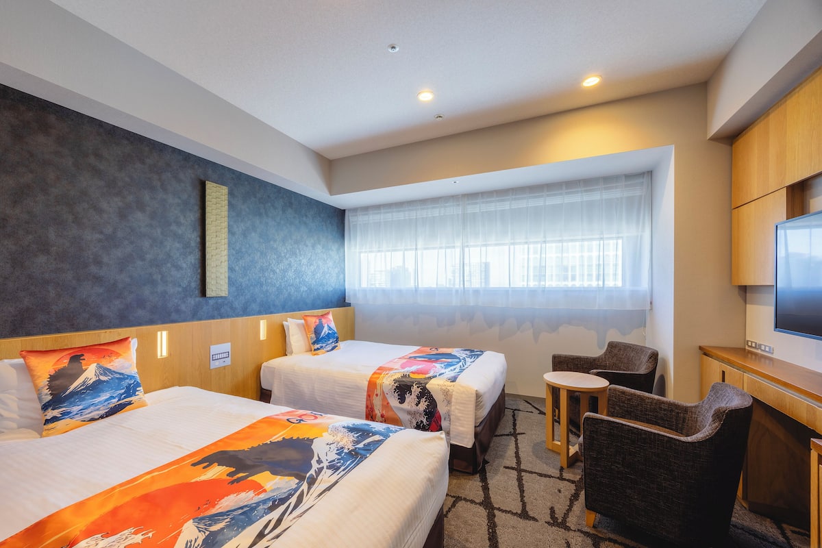 A hotel room with two beds, a desk, a chair, and a large window with sheer curtains.