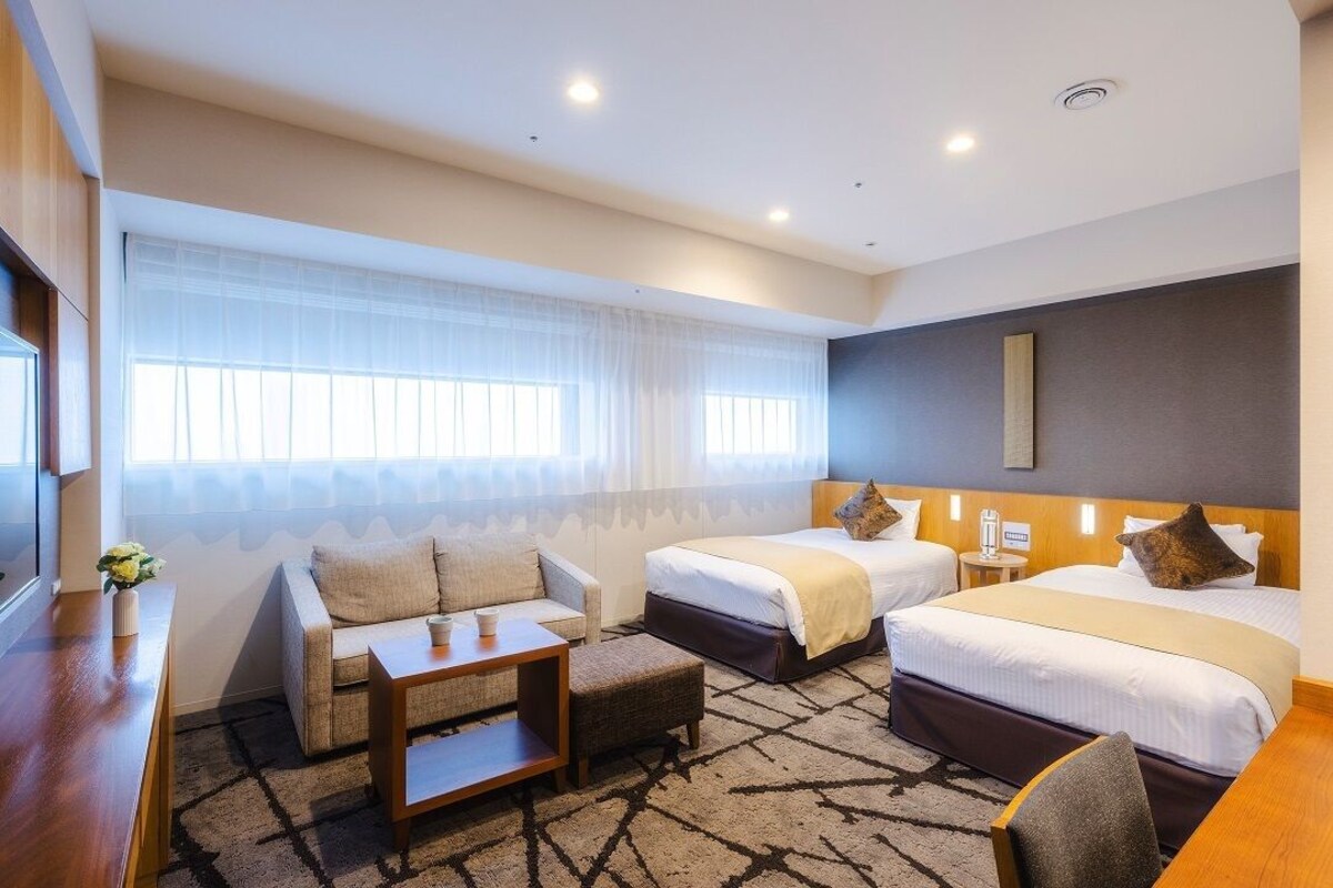 Habitación de hotel con dos camas, un sofá, una mesita y una ventana con cortinas translúcidas.