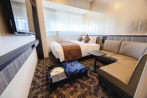 Una camera d'albergo con un letto, una poltrona, un tavolino e una valigia per terra.