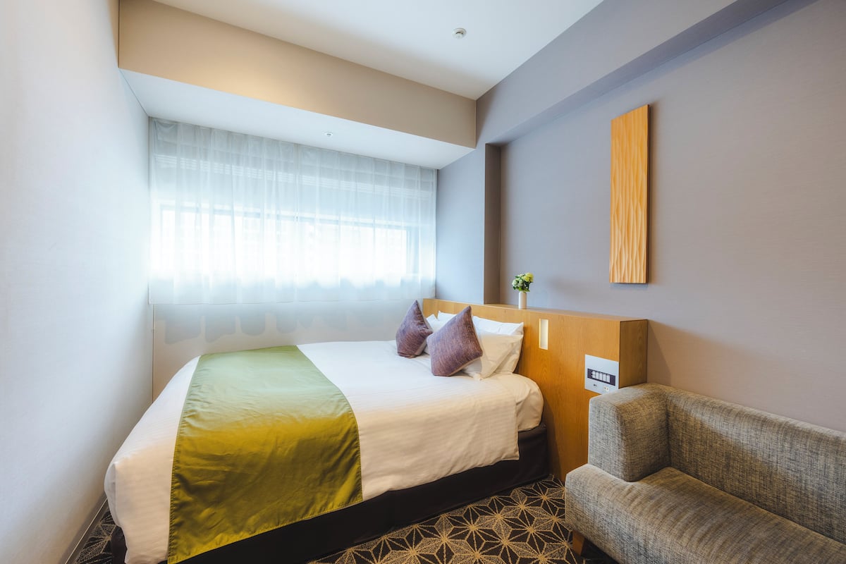 A hotel room with a bed, a chair, a window with sheer curtains, and a wall-mounted light fixture.